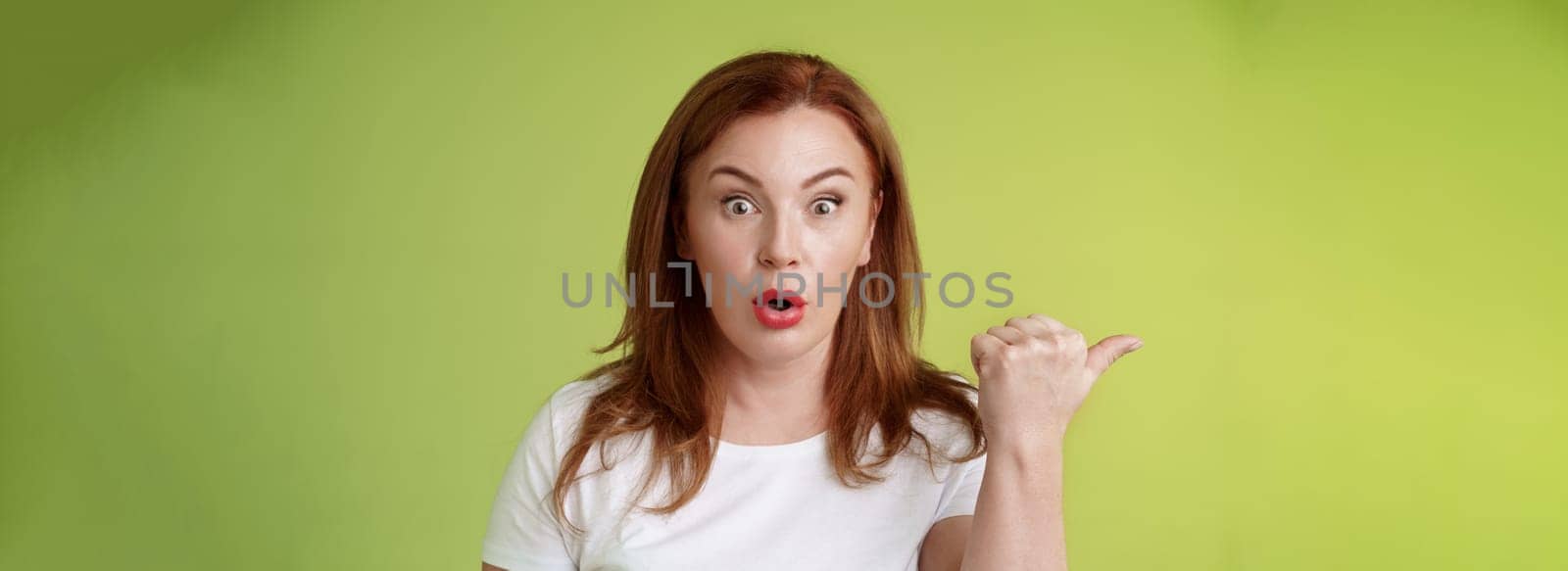 Close-up surprised amused redhead middle-aged housewife. folding lips say wow impressed stare camera questioned ambushed pointing left thumb astonished wondering if promo true green background.