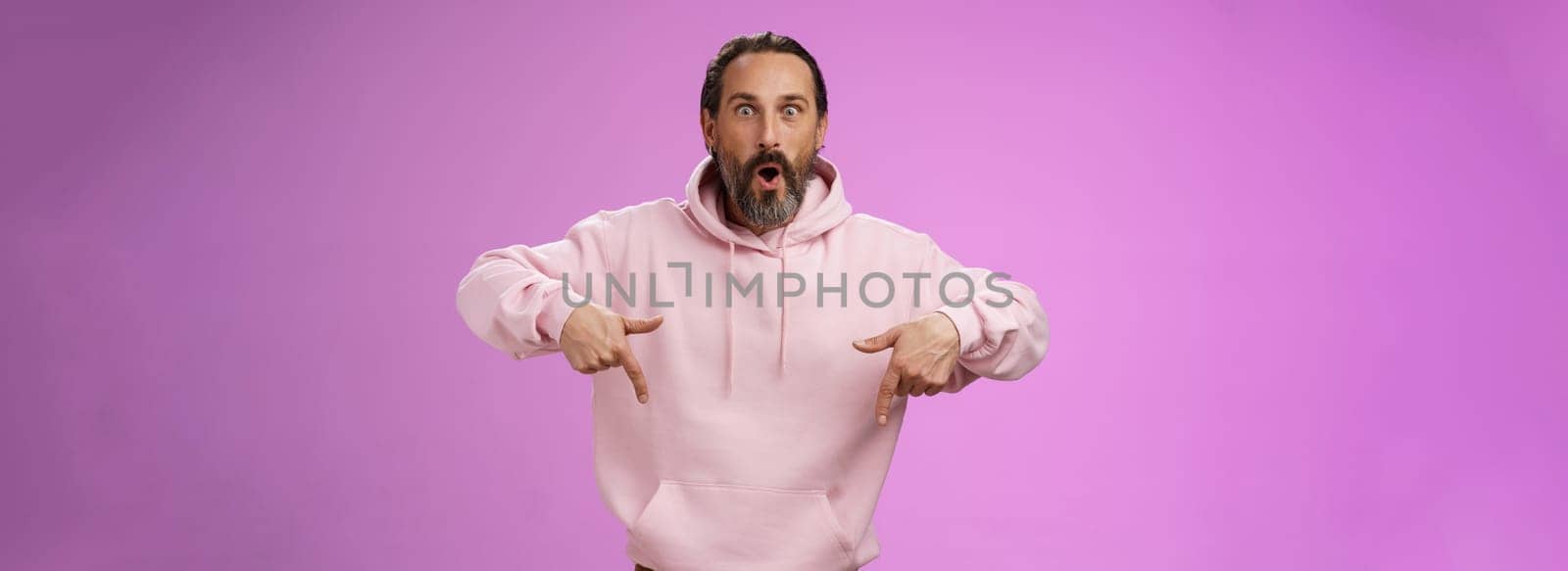 Impressed fascinated speechless dad bearded grey hair say wow widen eyes surprised folding lips amused curiously pointing down index fingers see super amazing advertisement, posing purple background.