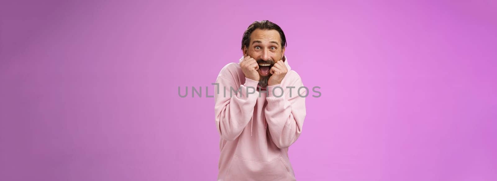 Amused thrilled gasping handsome happy bearded adult man in pink hoodie stooping screaming joyfully receive awesome incredible present standing speechless excited purple background. Copy space