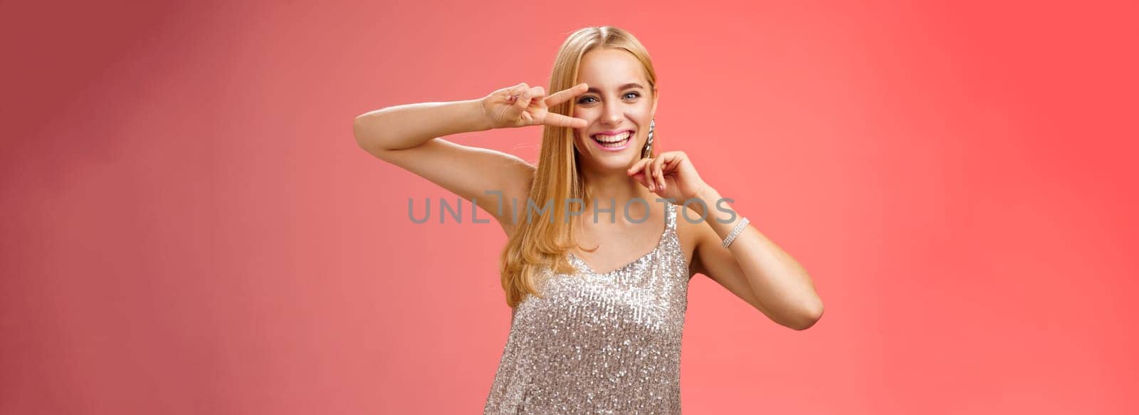 Charismatic funny amused charming blond girl 25s having fun dancing red background in silver trendy glittering party dress show peace disco gesture near eye smiling laughing carefree by Benzoix