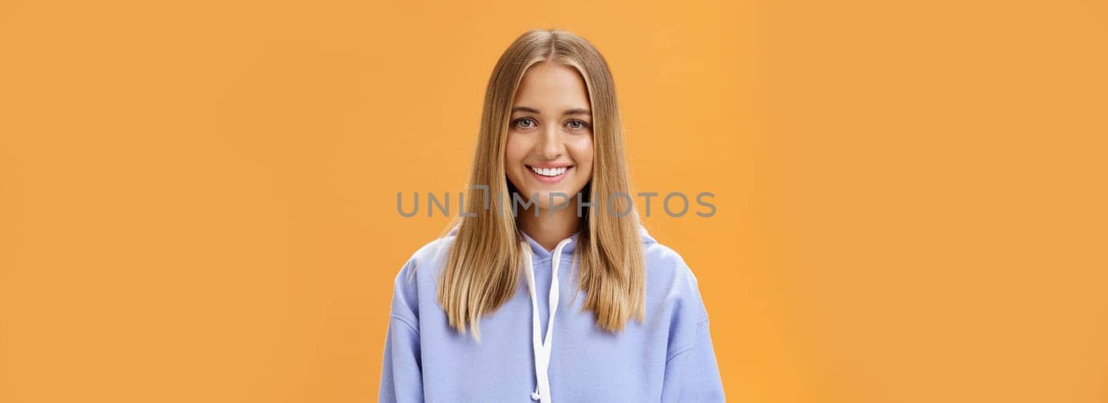 Waist-up shot of pleasant kind and happy friendly-looking young fair-haired girl in comfortable hoodie smiling cheerfully at camera with delighted carefree look posing against orange background. Advertisment and emotions concept