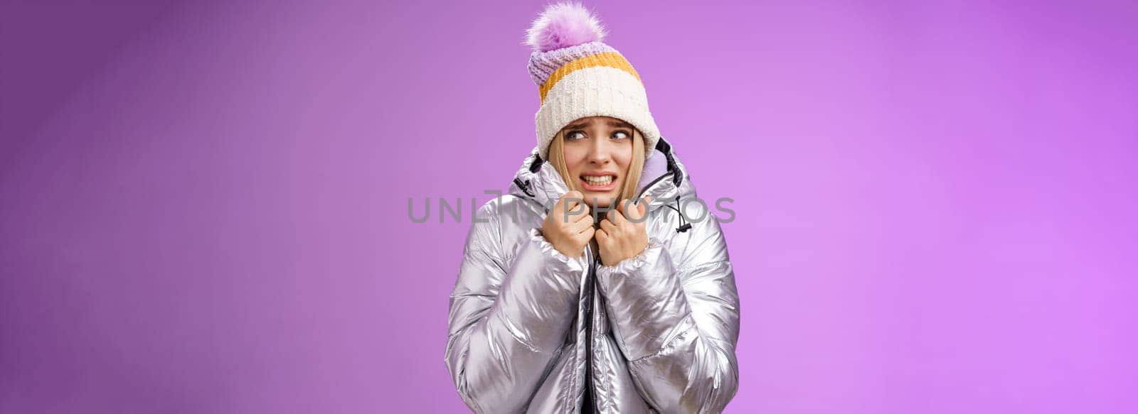 Freezing uncomfortable trembling cute blond girl in hat pull jacket tight feeling cold walking snowy mountains low temperature clenching teeth stooping shaking, standing upset purple background.