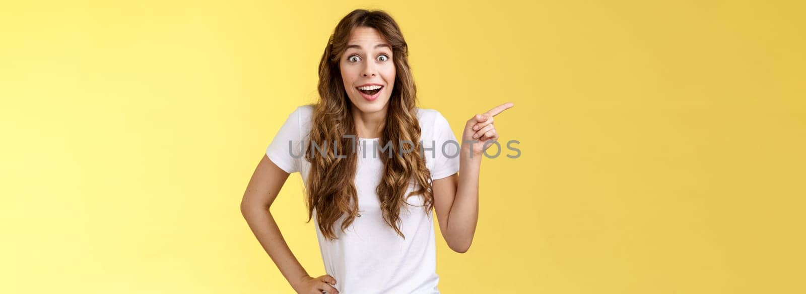 Surprised friendly sociable happy cute girlfriend long curly chestnut haircut smiling impressed gladly pointing left grinning camera toothy astonished expression discuss awesome performance.