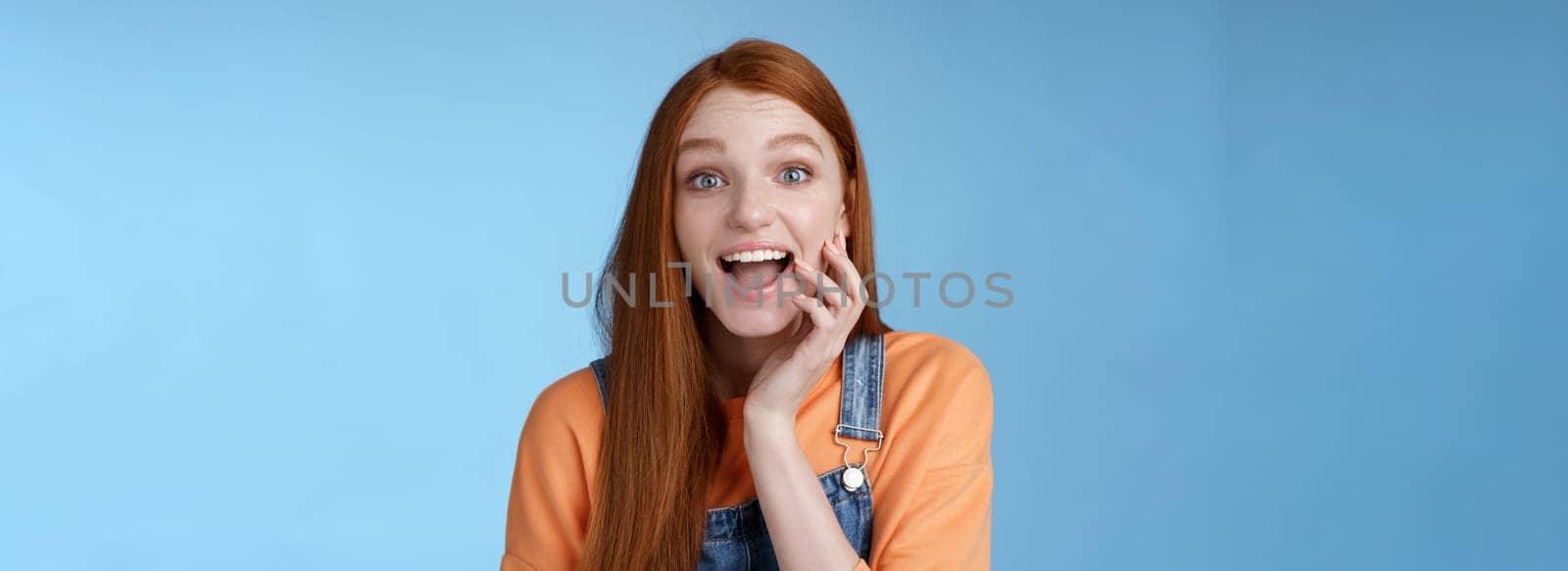 Excited enthusiasitc charismatic redhead female friend look surprised amused touch cheek open mouth thrilled discussing future prom rejoicing standing blue background joyful delighted.