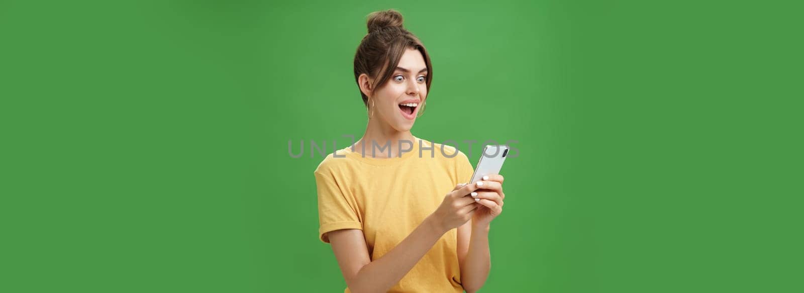Woman reading surprising satisfying message in smartphone opening mouth from excitement, smiling amazed looking astonished at cellphone screen posing against green background in casual yellow t-shirt. Technology concept