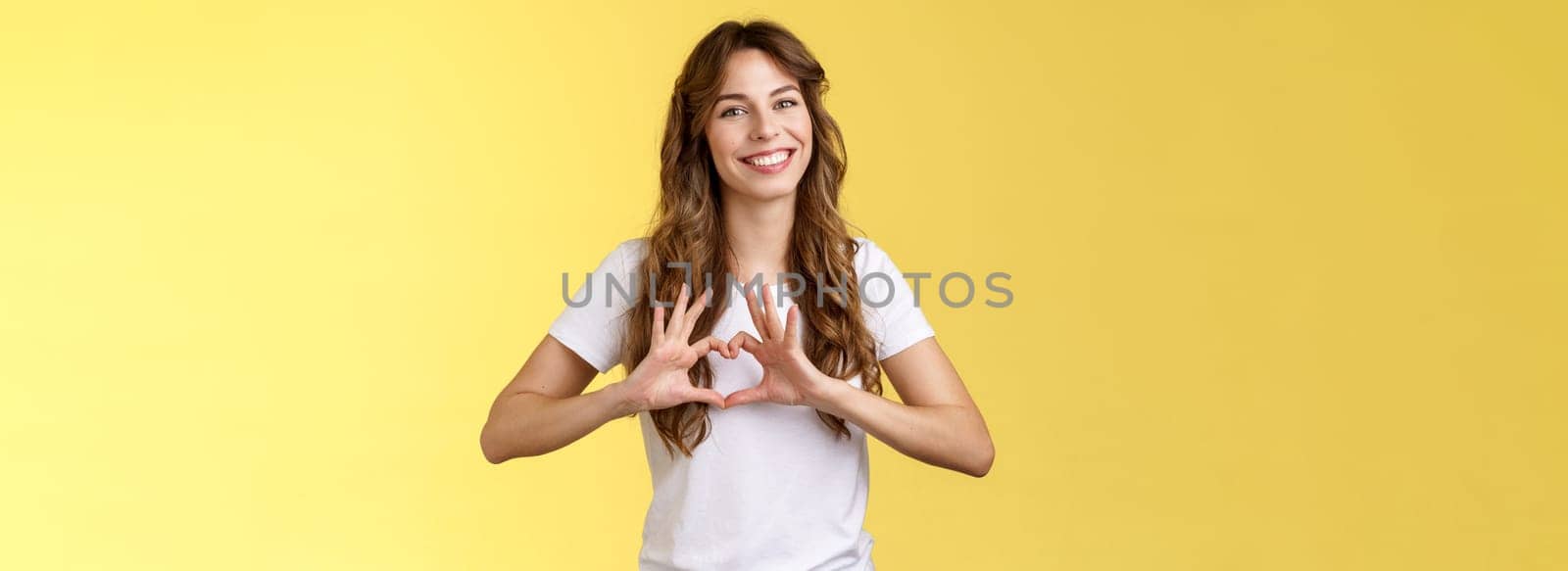 You my valantine. Tender confident adult girl curly hairstyle show heart gesture near chest express romantic sympathy feelings smiling delighted cherish relationship boyfriend yellow background.