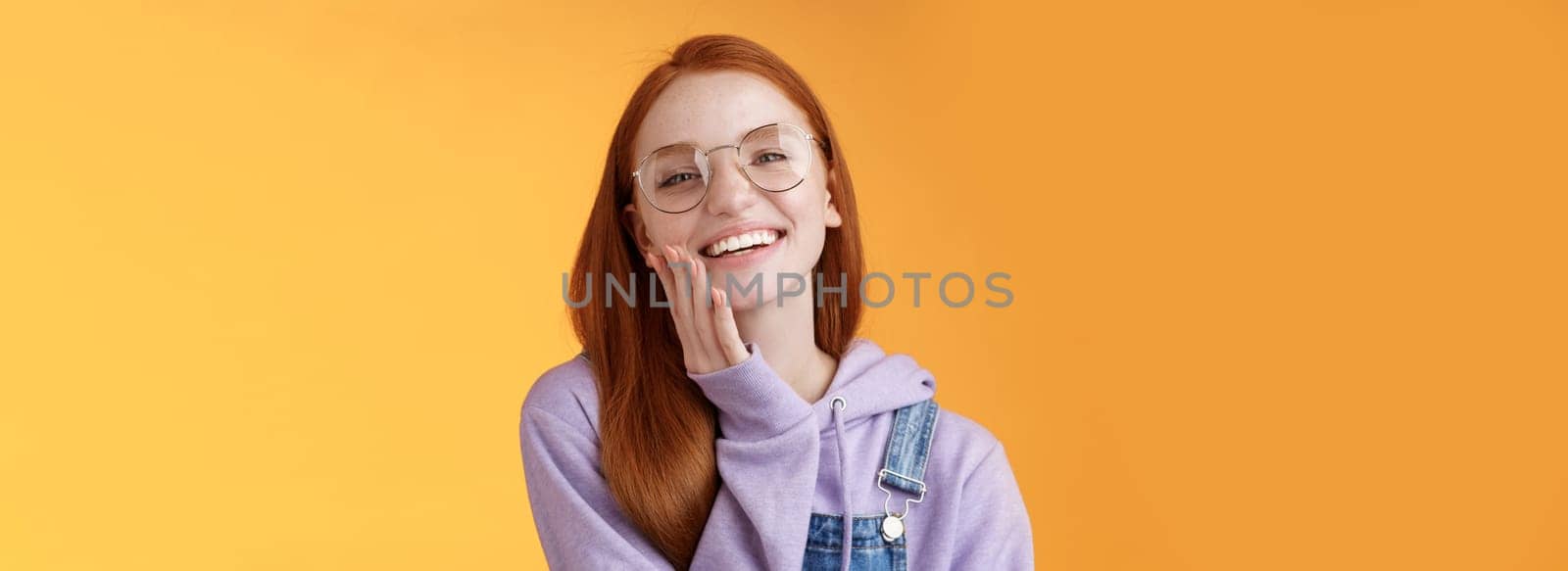 Charismatic happy tender redhead smiling european girl 20s glasses hoodie denim overalls having fun enjoying summer holidays chilly evening laughing joyfully touch pure clear skin, orange background.