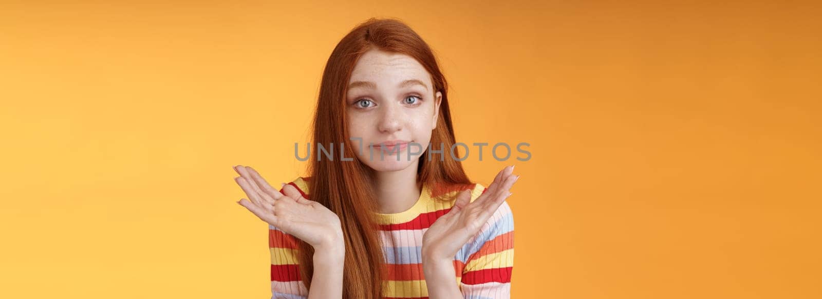 Clueless unbothered young redhead silly european girl 20s shrugging hands spread sideways smirking sorry cannot answer standing unaware confused puzzled give reply, orange background.