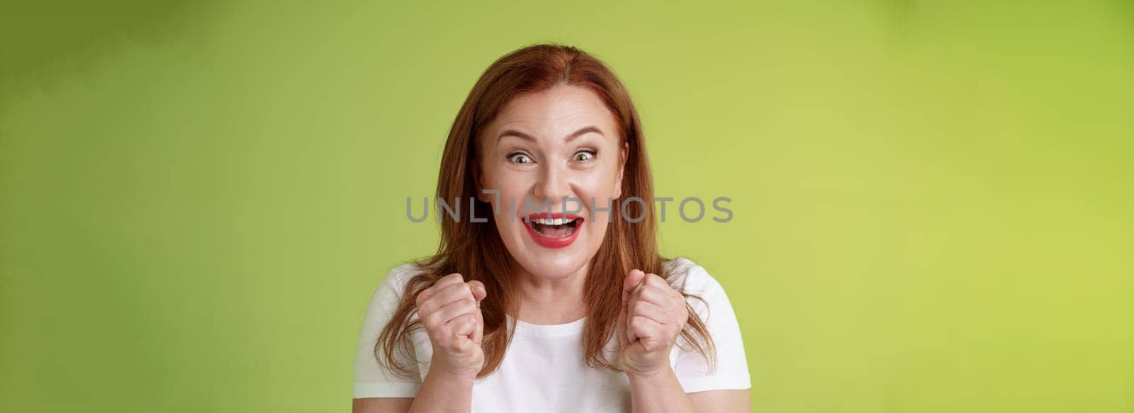 Close-up lucky enthusiastic cute redhead joyful middle-aged woman pump fists vigorous excitement celebratory smiling broadly winning celebrating triumphing success good news green background by Benzoix