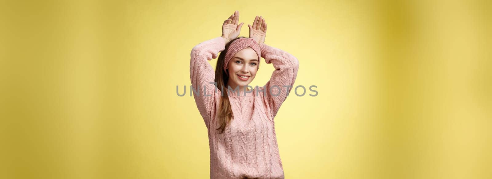 Nice charming playful young european female having fun, playing mimicking bunny showing rabbit ears with hands behind head, smiling cute, tender, happy greeting boyfriend welcome back ove yellow wall by Benzoix
