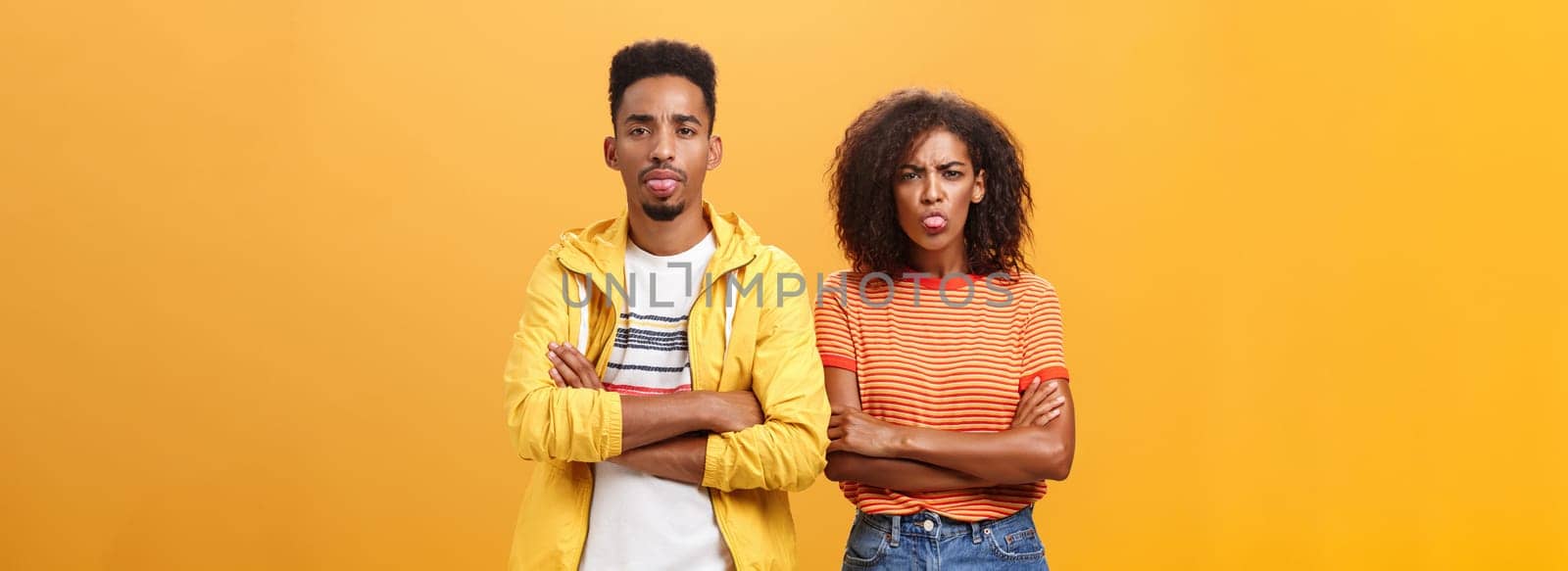 Indoor shot of african american siblings being displeased and annoyed showing bad tempber behaving childish sticking out tongue standing together with crossed hands on chest over orange background. Lifestyle.