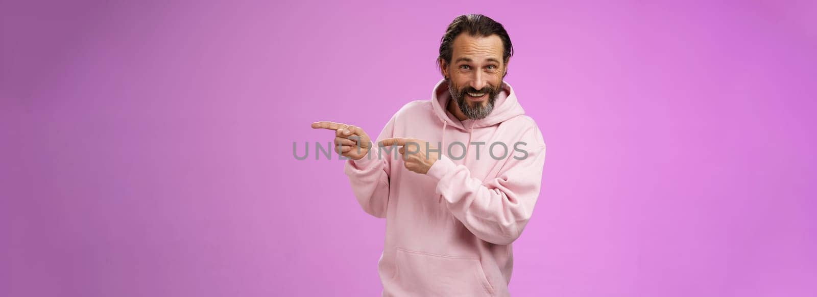 Sassy confident good-looking macho man 50s in pink hoodie smiling broadly inviting join pointing right showing interesting amusing place hang out welcoming take look, standing purple background.