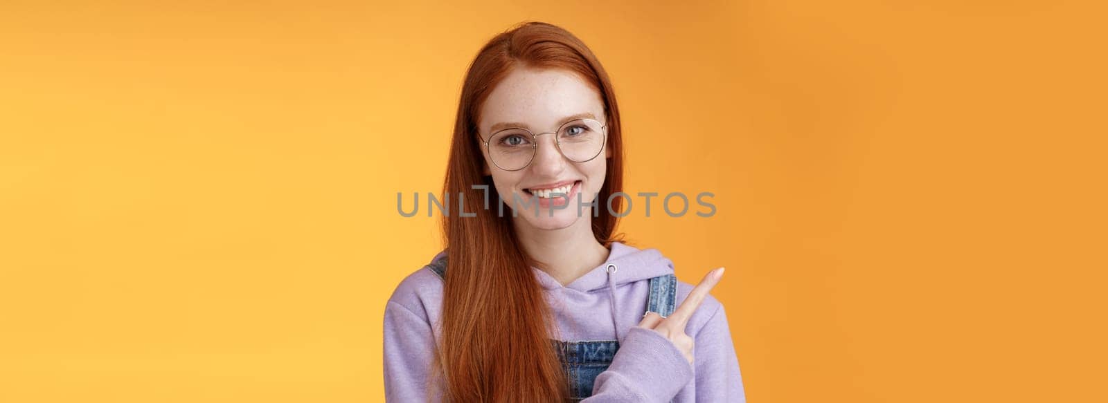 Friendly good-looking modern redhead young girl pointing left index finger showing awesome place suggesting go hang out smiling joyfully casually talking discuss new product, orange background by Benzoix