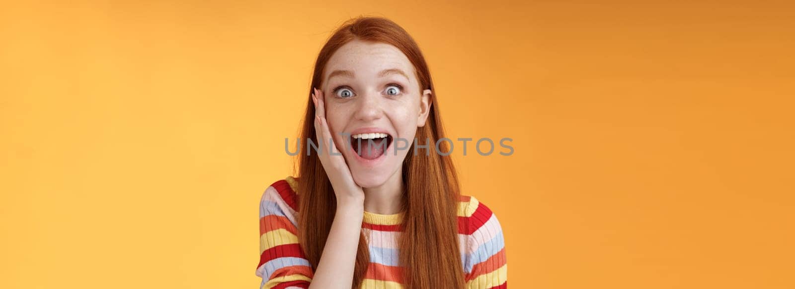 Amused happy smiling good-looking redhead woman screaming happily touch cheek astonished receive good perfect news triumphing feeling excited look thrilled camera, standing orange background.