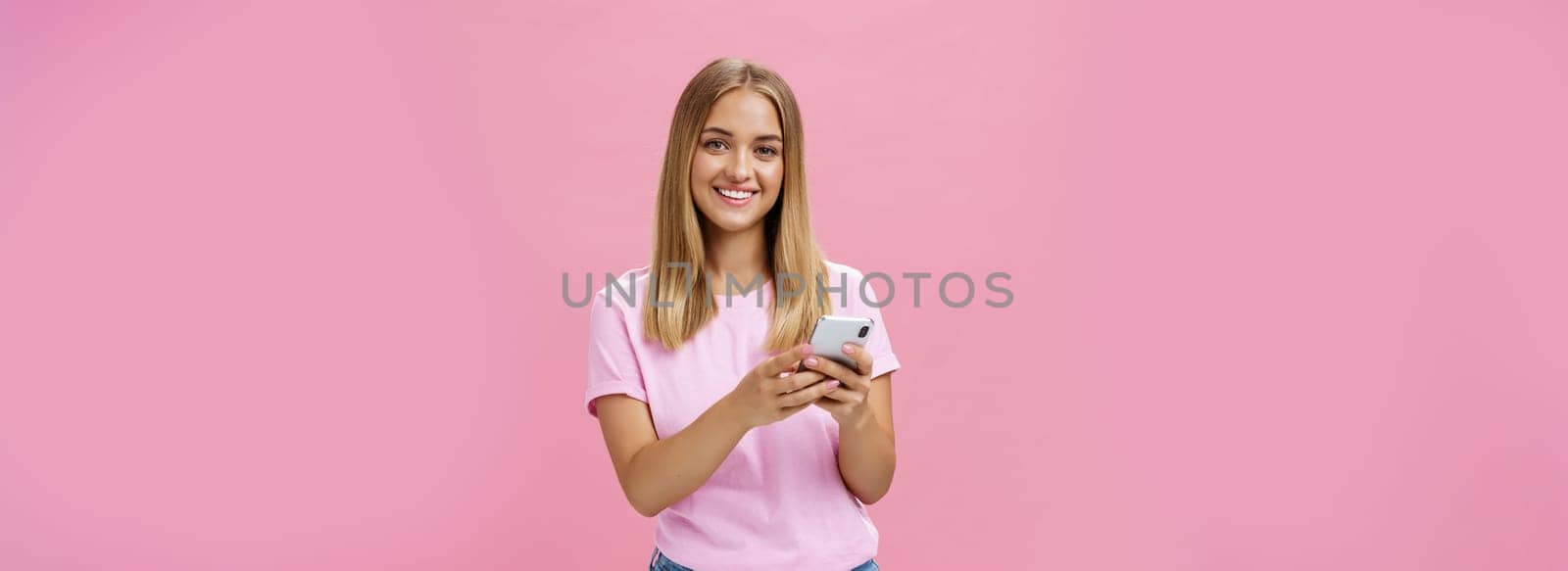 Woman calling taxy via smartphone asking friend address smiling cheerfully at camera holding phone with both hands over chest getting in touch with clients via messages standing over pink wall. Technology concept
