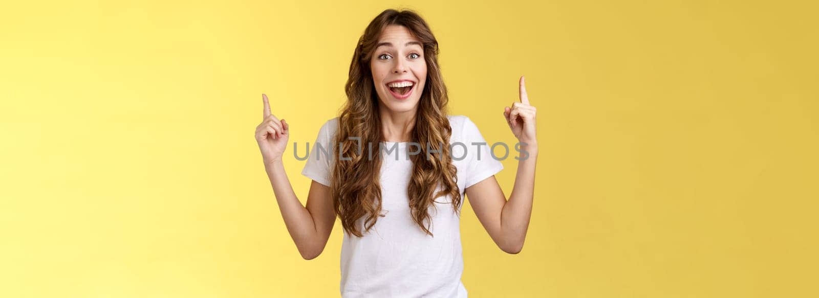 Surprised cheerful celebrating girl lively discuss near awesome event pointing up index fingers smiling broadly fascinated look temtation admire great concert stand yellow background impressed.