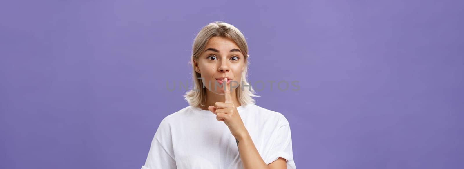 Lifestyle. Charming creative and stylish beautiful tanned girl with fair hair shushing at camera with delighted thrilled look making up great idea wanting hide it and keep secret posing over purple wall.