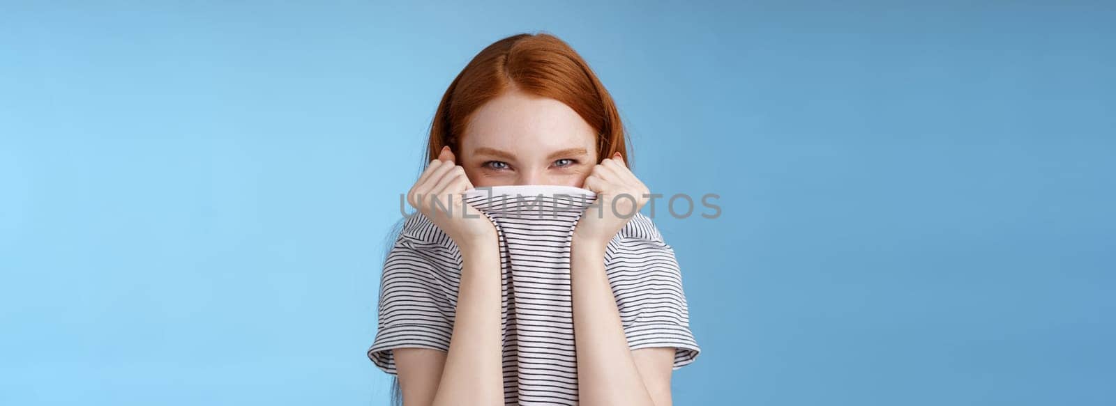 Silly flirty amused attractive playful redhead girlfriend hiding face pulling t-shirt head squinting devious mysteriously giggle laughing hope disguise pranking friend standing blue background.