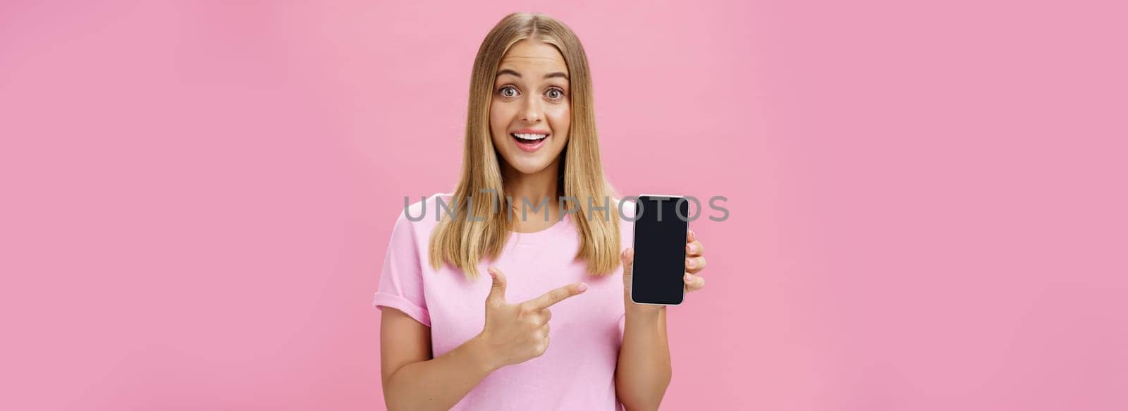 Cheerful attractive and pleasant woman promoting cool app or smartphone holding cellphone and pointing at device screen smiling amused and impressed standing over pink background by Benzoix