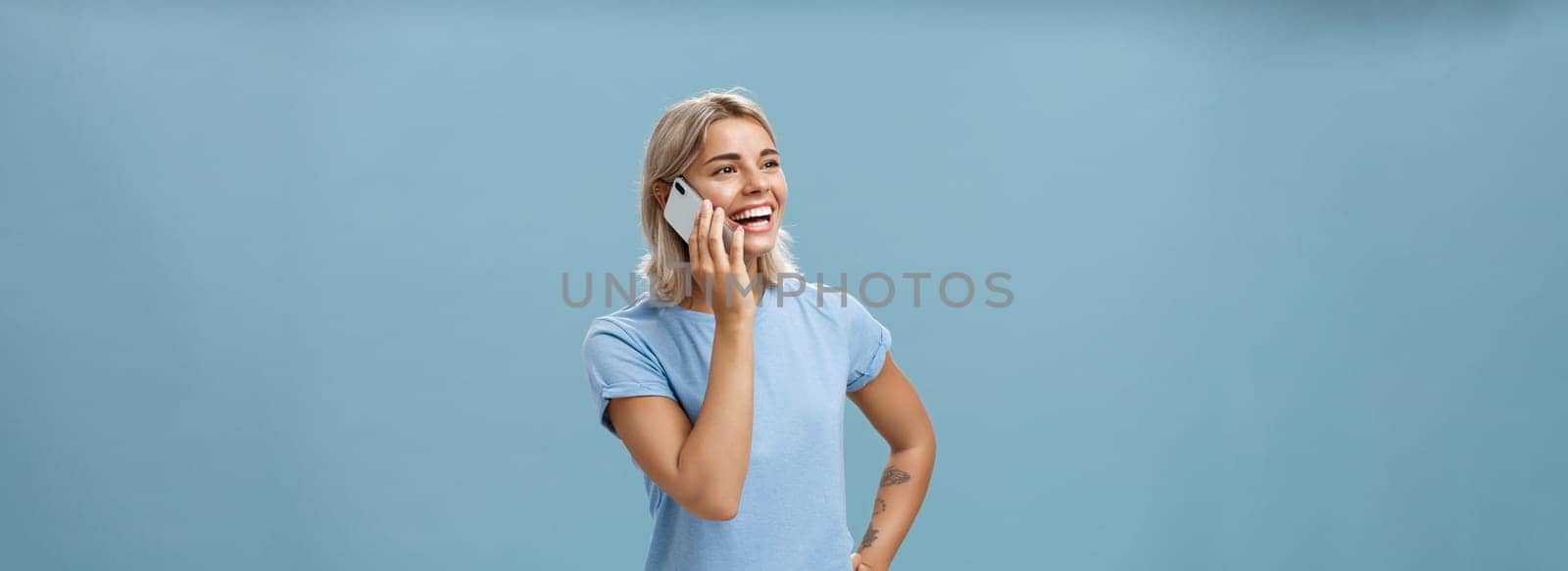 Waist-up shot of sociable amused and happy attractive caucasian fair-haired woman in casual t-shirt standing half-turned gazing left with hand on hip while talking on smartphone over blue background by Benzoix