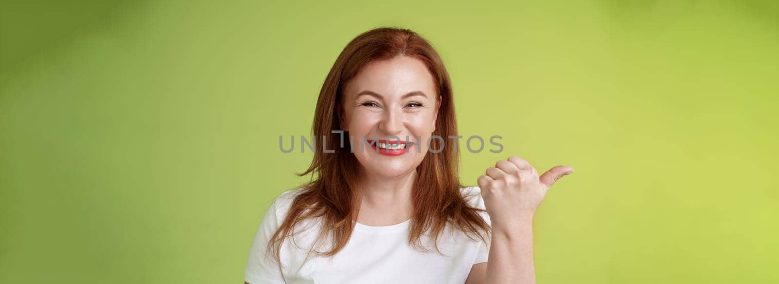 Come visit our store. Cheerful pleasant friendly charming redhead middle-aged woman entrepreneur inviting check-out promo smiling happily sincere kind grin pointing left thumb stand green background.
