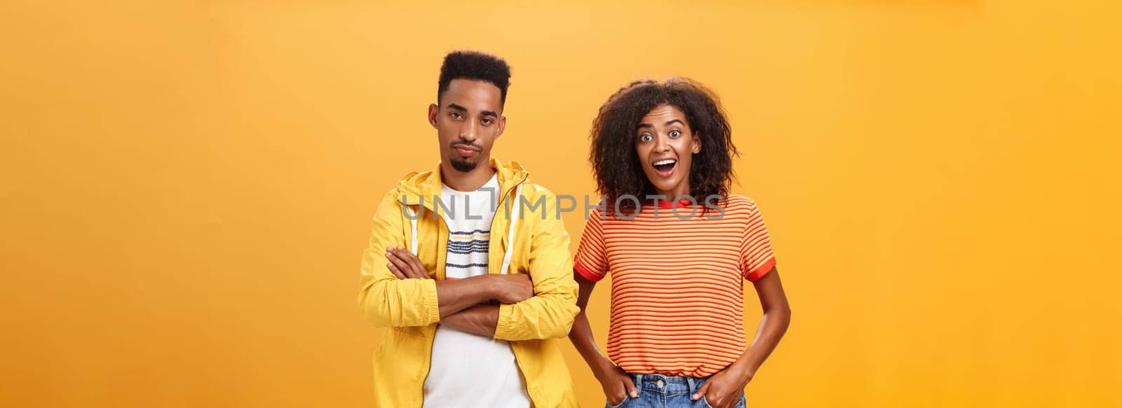 Two african american friends standing over orange background, girl thinks idea is awesome feeling excited and joyful while boyfriend being displeased and indifferent to unimpressive plan by Benzoix