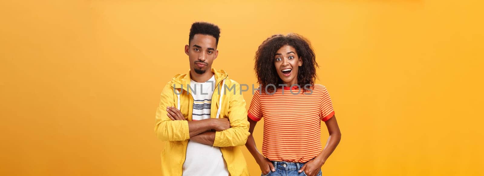 Two african american friends standing over orange background, girl thinks idea is awesome feeling excited and joyful while boyfriend being displeased and indifferent to unimpressive plan. Copy space