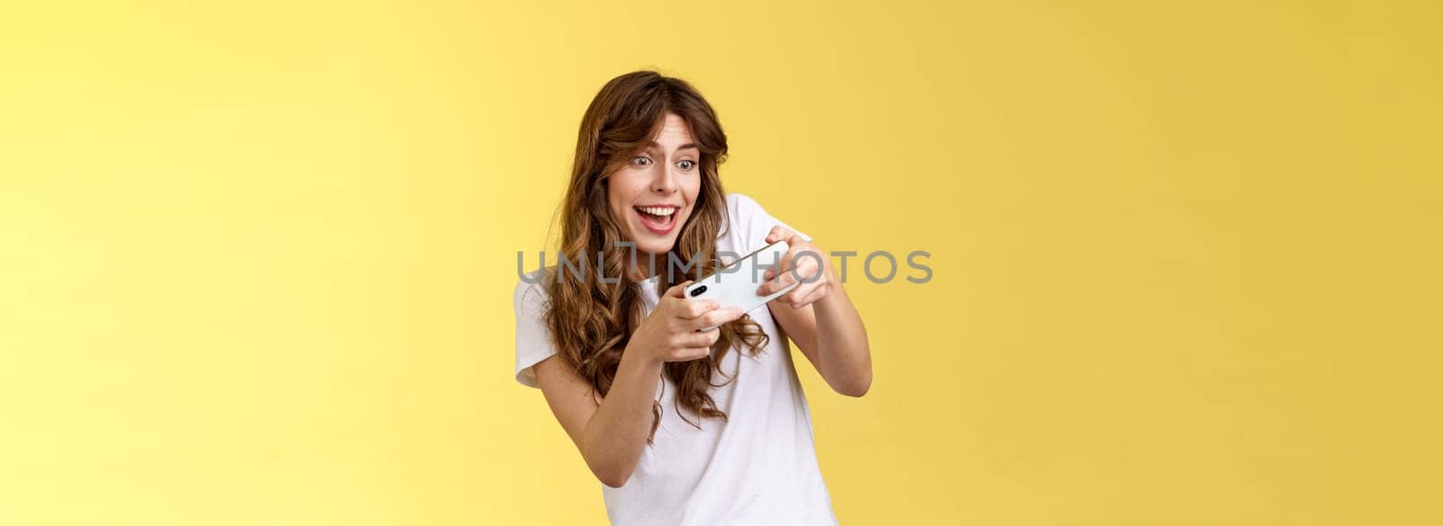 Excited playful enthusiastic girl tilting sideways playing awesome interesting smartphone game car racing smiling determined focuse gaming hold mobile phone horizontal tap display yellow background.