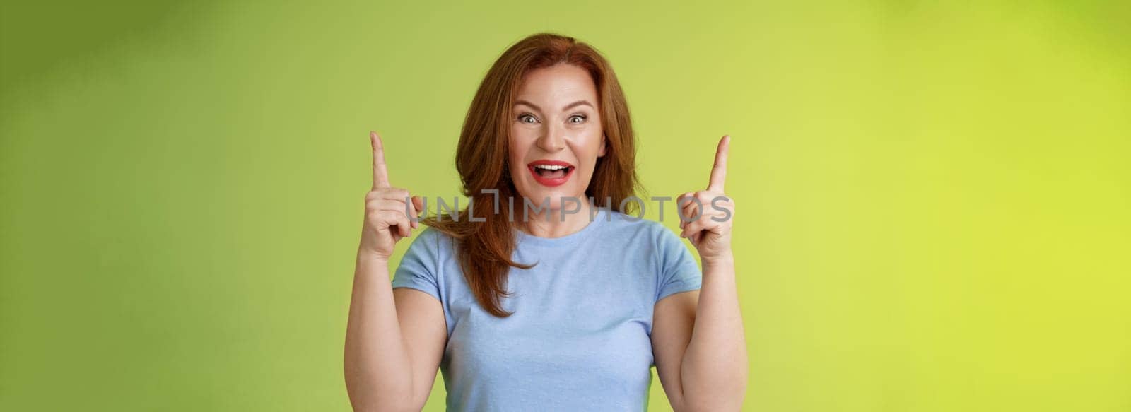 Impressed enthusiastic happy cheerful middle-aged mature woman red hair smiling astonished look admiration joy pointing up raised index fingers introduce impressive stunning offer green background by Benzoix