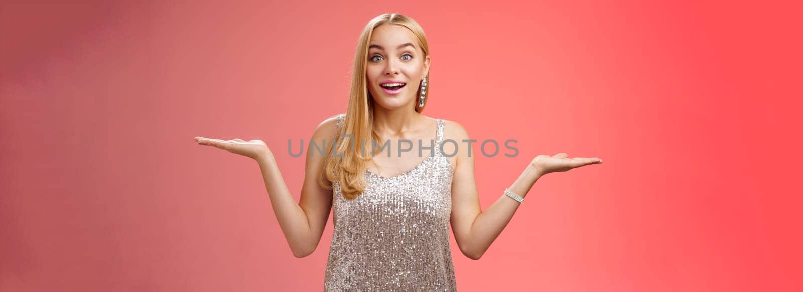 Carefree fascinated young blond girl 20s in silver glittering evening party dress raising hands sideways weighing shrugging smiling amused search right choice, deciding what do red background.