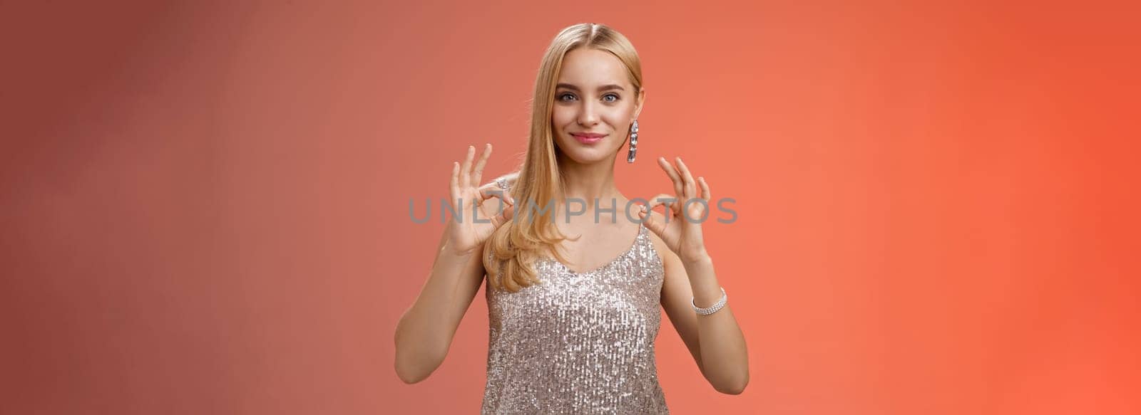 Confident carefree optimistic young blond woman assure everything done perfect liking result show okay ok no problem gesture smiling self-assured standing silver luxurious dress, red background.