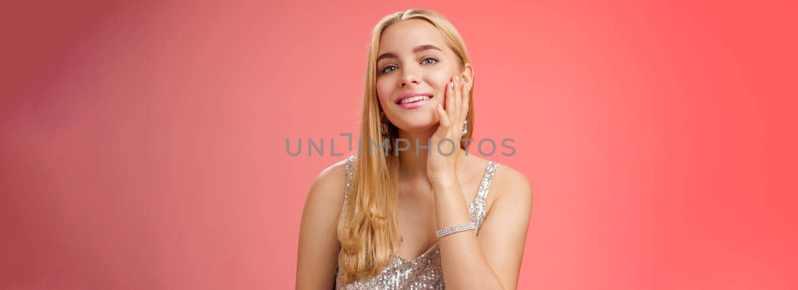 Tender stylish self-assured coquettish young blond wealthy woman in silver shiny dress touching pure clean skin satisfied good makeup get ready party smiling delighted, standing red background.