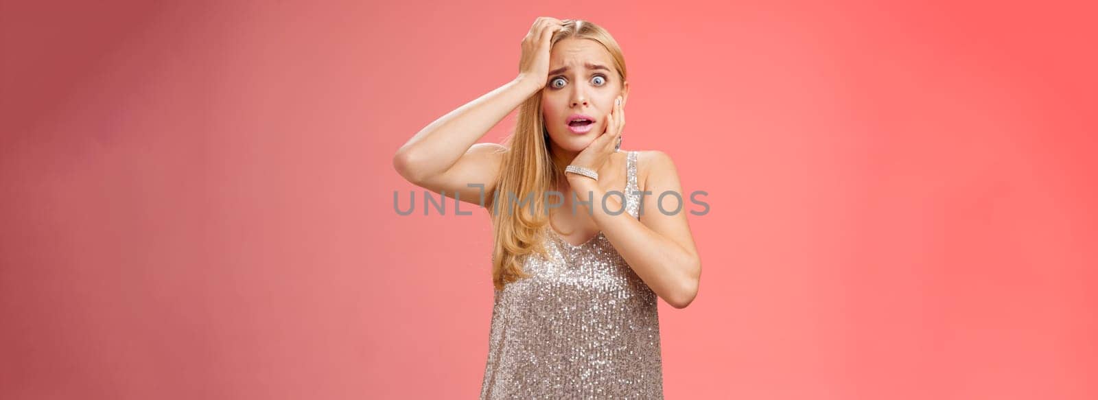 Shocked frightened insecure panicking woman blond hairstyle in silver dress touch head pop eyes afraid scared seing terrifying crime standing stupor speechless gasping shook, red background.