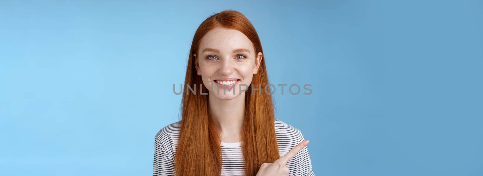 Pleasant charismatic helpful friendly-looking female redhead blue eyes pointing left index finger help make decision pick product store smiling broadly confident gaze camera asking trust her by Benzoix