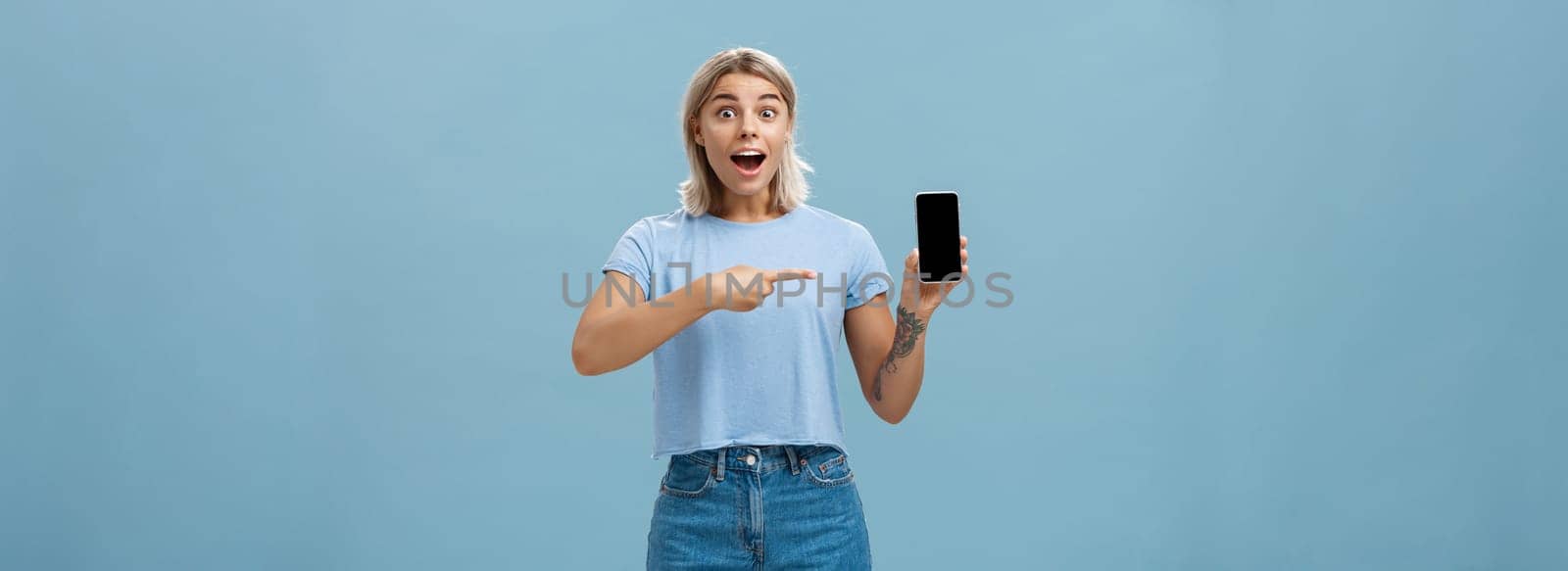 Studio shot of amazed and impressed charming european blonde in casual clothes gasping charmed at camera while showing smartphone screen pointing at device with finger over blue background by Benzoix