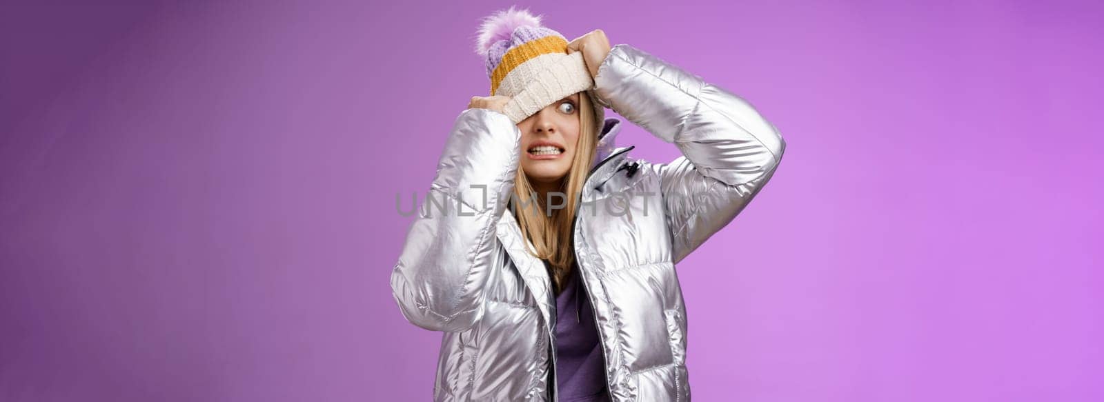 Awkward embarrassed cute blond girl worried pulling hat face hiding from ex-boyfriend pretend not see him standing anxiously clenching teeth peeking one eye left nervous, purple background by Benzoix