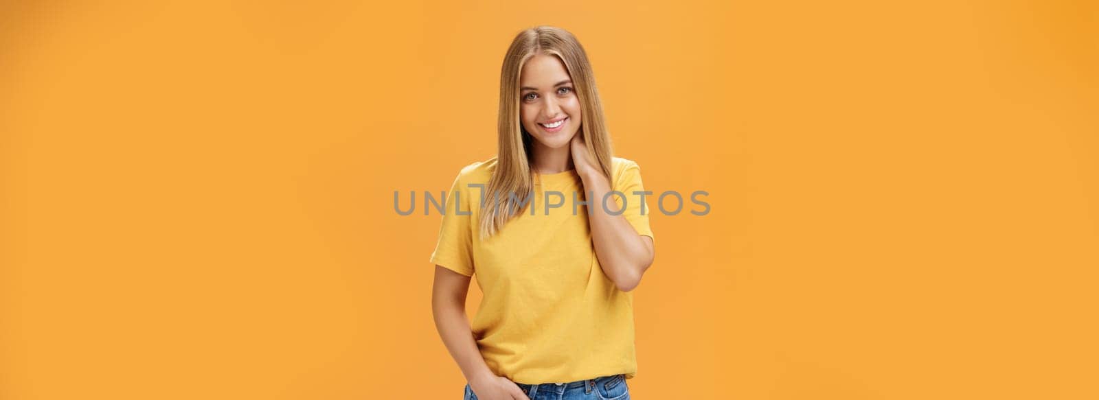 Portrait of shy and timid feminine girl with tan and straight fait hair rubbing neck and smiling sensually with happy carefree expression holding hand in pocket posing against orange background. Lifestyle.