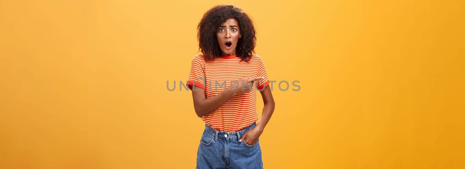 Studio shot of offended shocked stylish woman being robbed fasping and staring nervously begging stranger help out pointing left concerned and worried frowning, posing displeased over orange wall by Benzoix