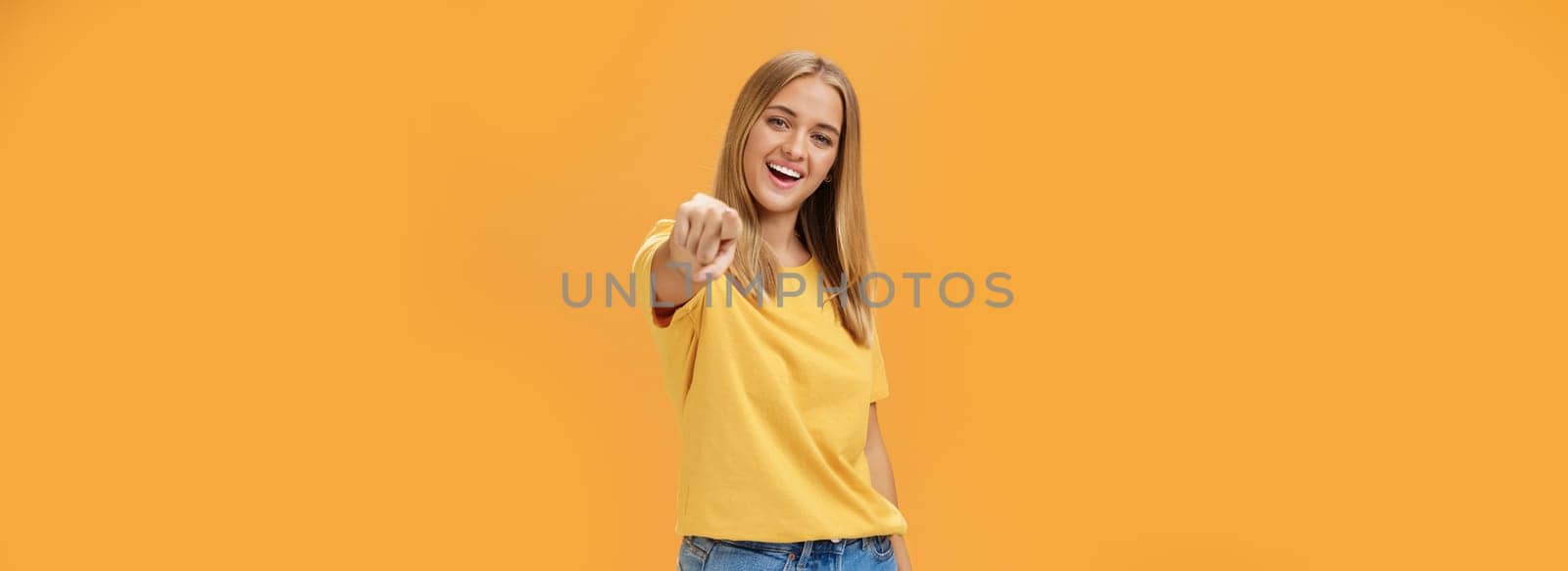 Woman directing at camera with index finger and smiling friendly as if talking to us standing amused and happy with confident carefree expression pulling arm towards to point over orange background by Benzoix