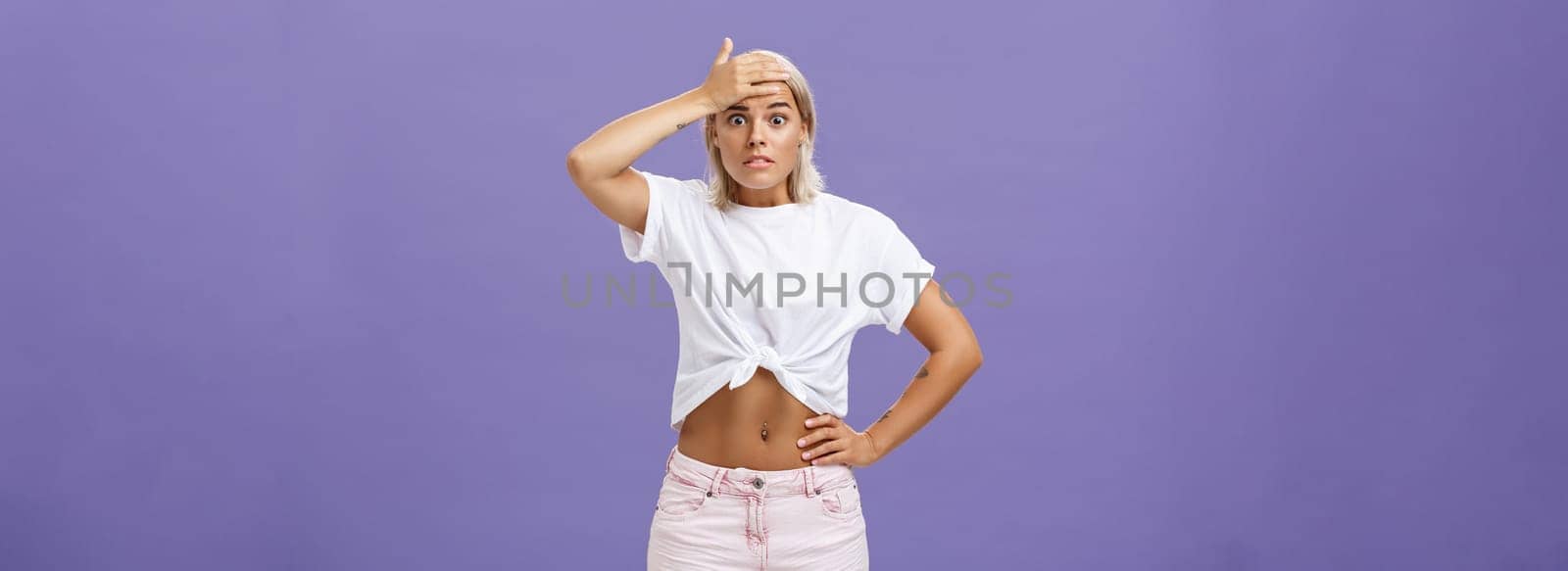 Damn I am in trouble. Worried troubled good-looking stylish female employee with tanned skin and fit body holding hand on forehead popping eyes from anxiety and making nervous face over purple wall by Benzoix