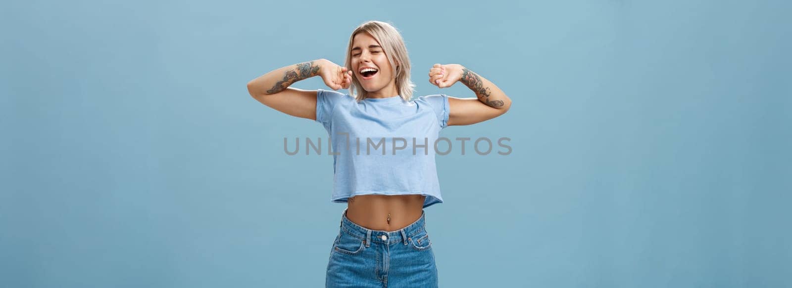 Great feelings after nap. Satisfied carefree and relaxed attractive athletic and stylish female with tattooed arms stretching hands yawning and smiling with closed eyes and pleased look after sleep. Emotions concept