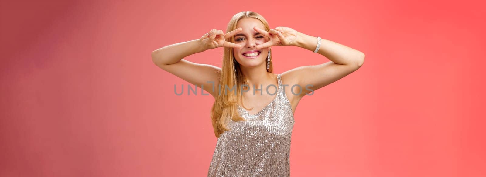 Sassy attractive stylish european girlfriend having fun celebrating birthday standing silver evening glittering dress winking cheeky make disco peace gesture near eyes enjoying party, red background.