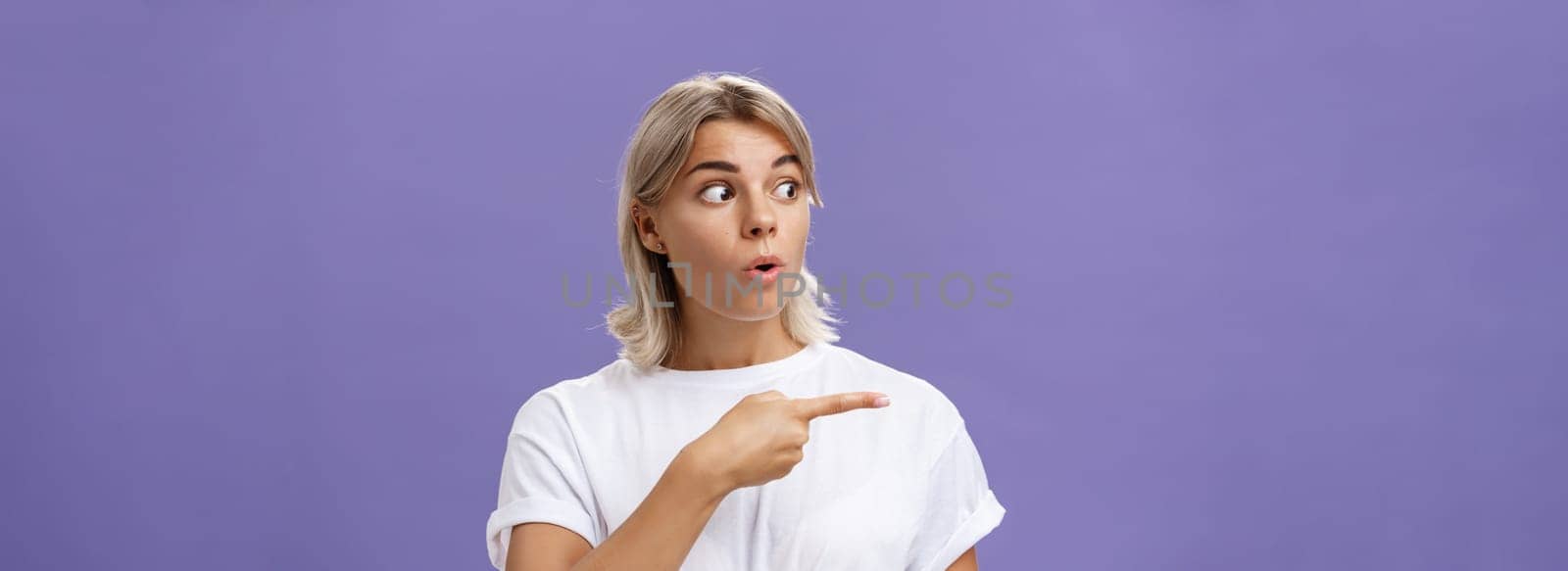 Close-up shot of intrigued interested attractive adult blonde female in white t-shirt folding lips staring and pointing left with curious and thrilled expression over purple background by Benzoix