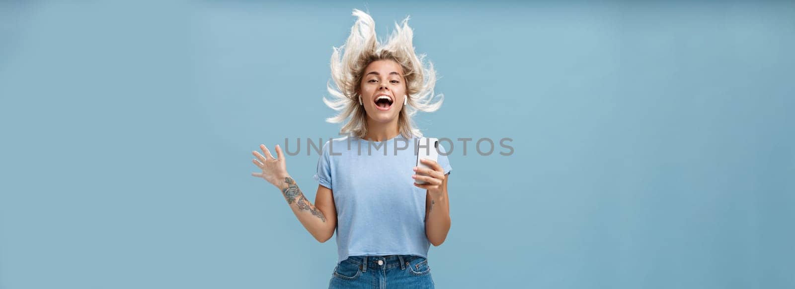 Expressing happiness with help of great tunes. Joyful amused and happy good-looking young female student jumping having fun listenign music in wireless earbuds, holding smartphone over blue wall by Benzoix