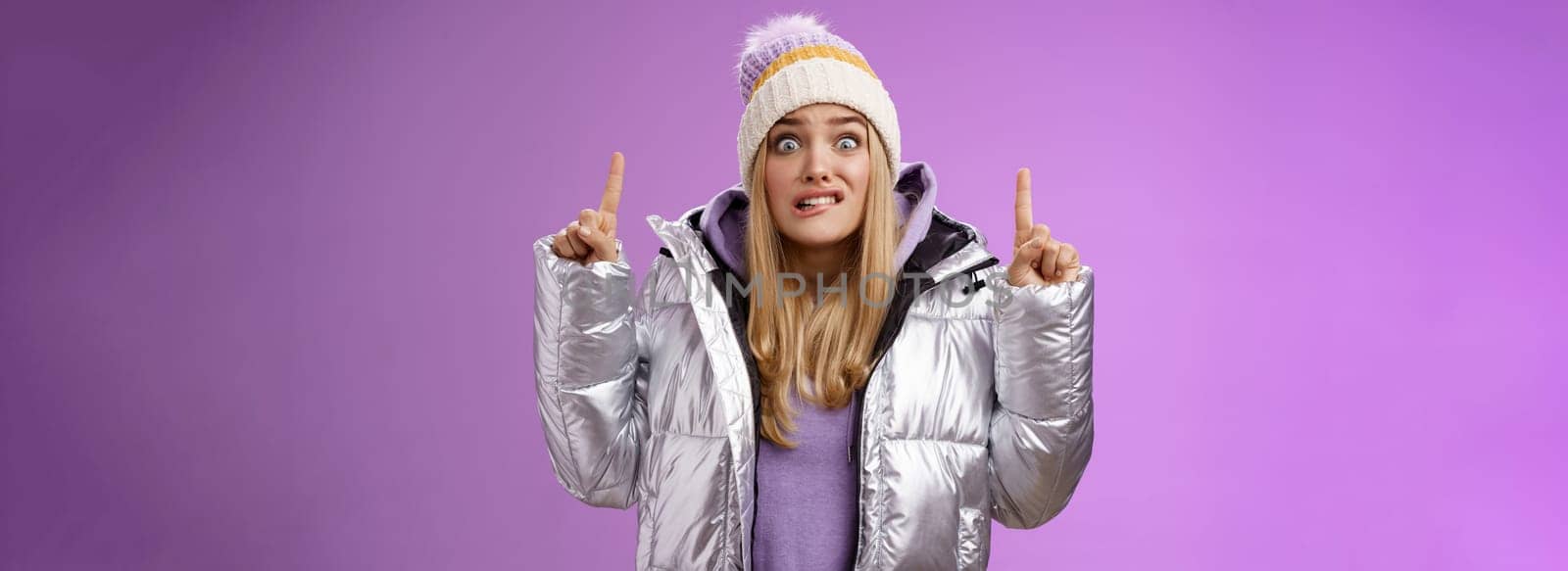 Scared unsure worried awkward young cute blond girl biting lip stooping pointing up widen eyes nervously scared someone find out secret standing embarrassed and intense purple background by Benzoix