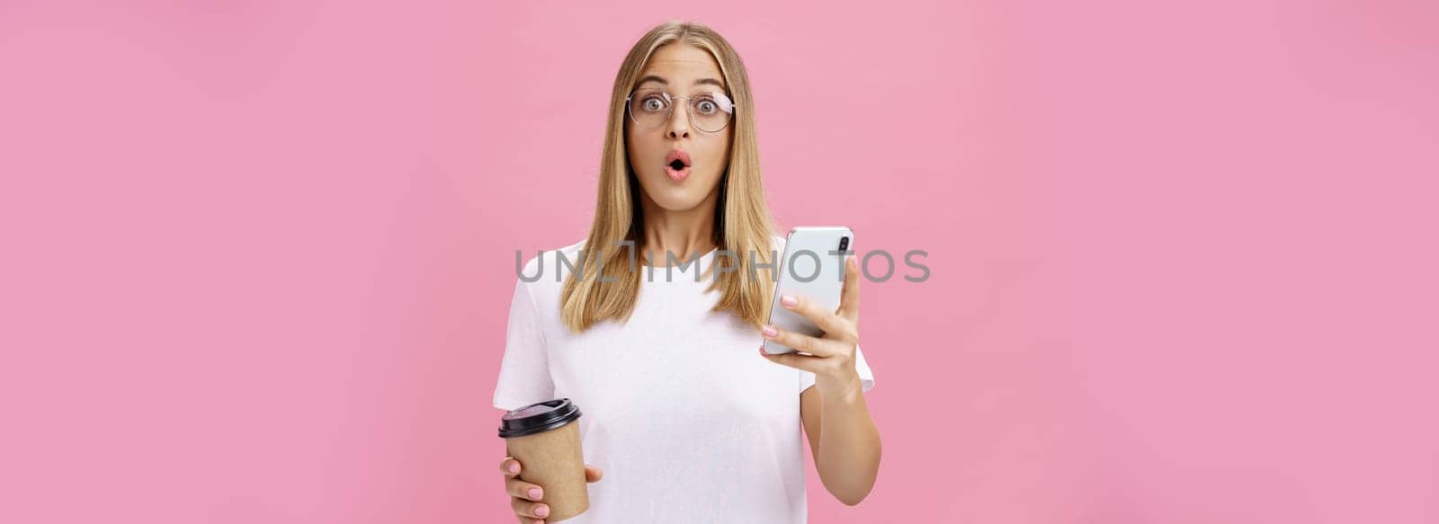 Girl receiving awesome message on smartphone while drinking coffee from cup in cafe sharing news with friend folding lips in wow sound staring surprised at camera holding cellphone over pink wall by Benzoix