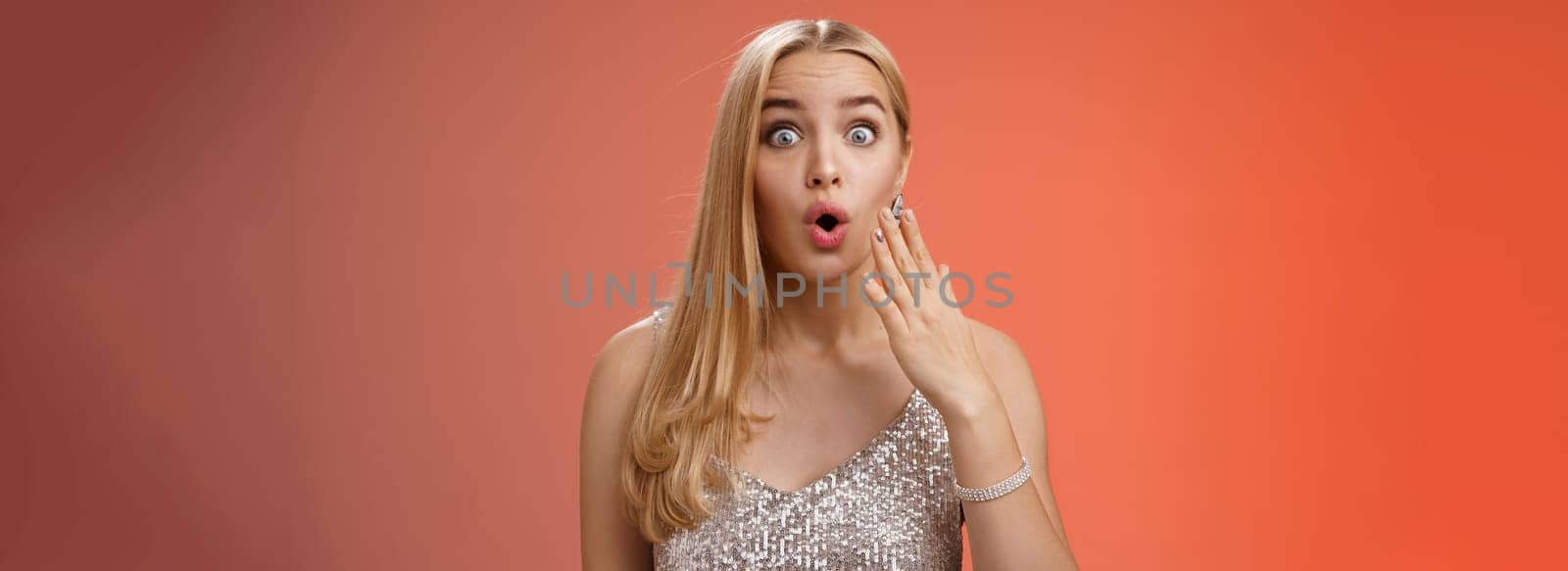 Amused wondered good-looking elegant blonde in silver stylish dress pop eyes surprised folding lips wow cover mouth palm amazement learn incredible rumor gossip with friend, red background.