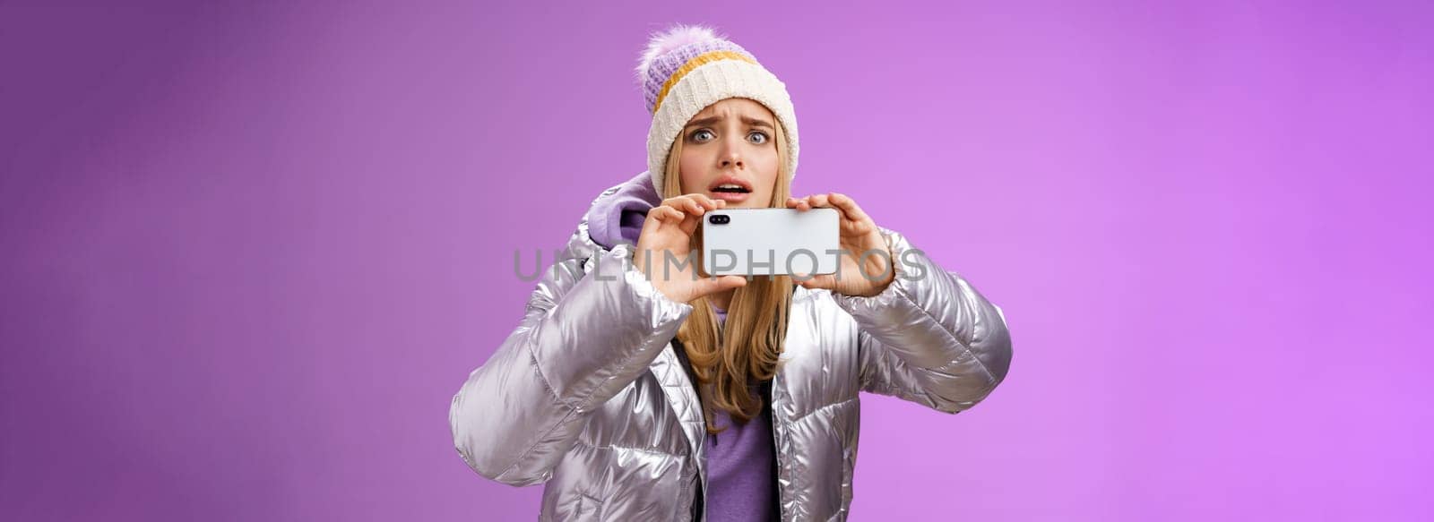 Intense worried blond girl holding mobile phone horizontal recording video capture moment share friends internet blog using smartphone camera shoot photograph standing serious purple background.