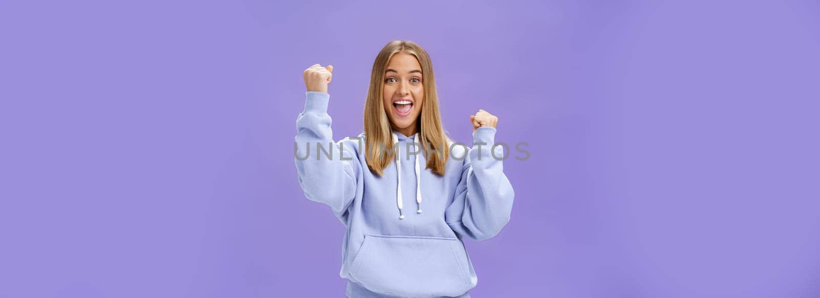 Cheerful happy and supportive young girlfriend with fair hair and tan in warm hoodide raising fists in cheer and triumph smiling saying yeah celebrating goal or success over purple background. Lifestyle.
