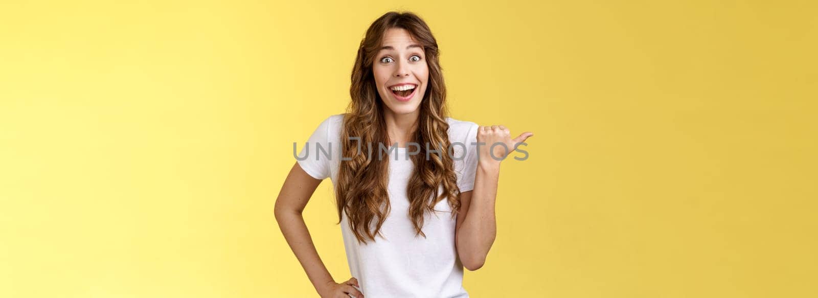 So amazing lets go together. Lively excited european female curly haircut wanna visit cool party smiling joyfully pointing thumb left grinning thrilled look camera admiration yellow background.